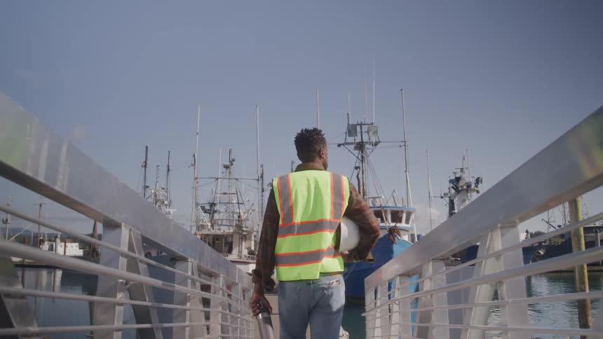 A man is standing with his back turned and walking on a bridge to a marina full of boats and he is wearing a green vest with yellow lines