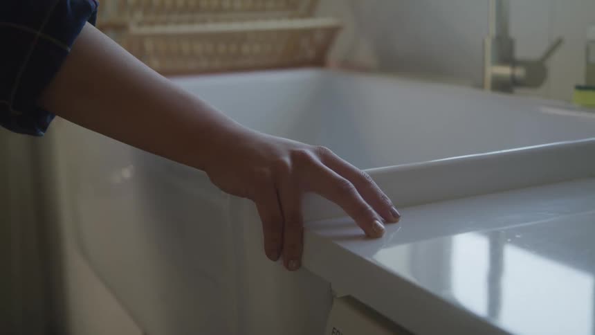 A persons hand on the corner of a white sink that is reflecting the environment