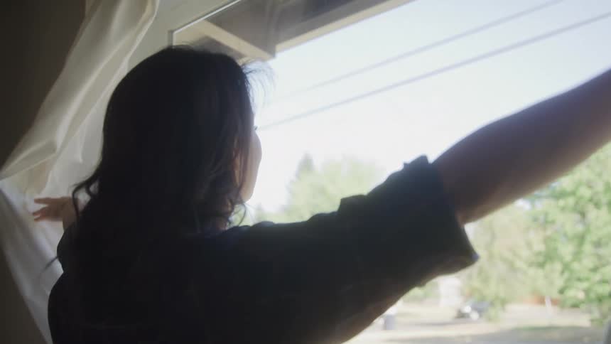A woman pulling the drapes and looking out the window into the yard