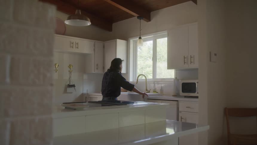 A woman leans against the sink and looks out the window