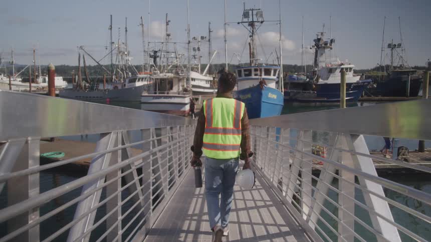 A man in a green vest is walking backwards on a metal walkway going to a pier