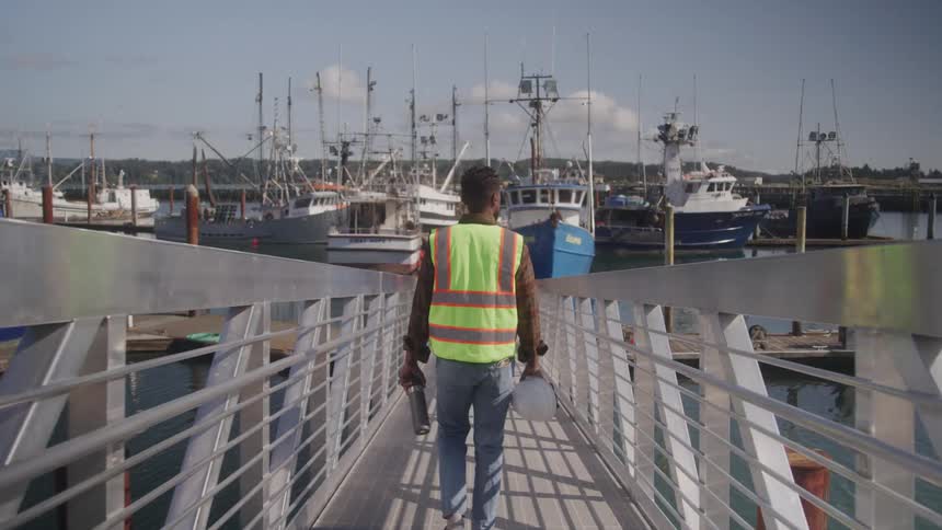 A worker in a reflective vest walks toward fishing boats at a coastal dock