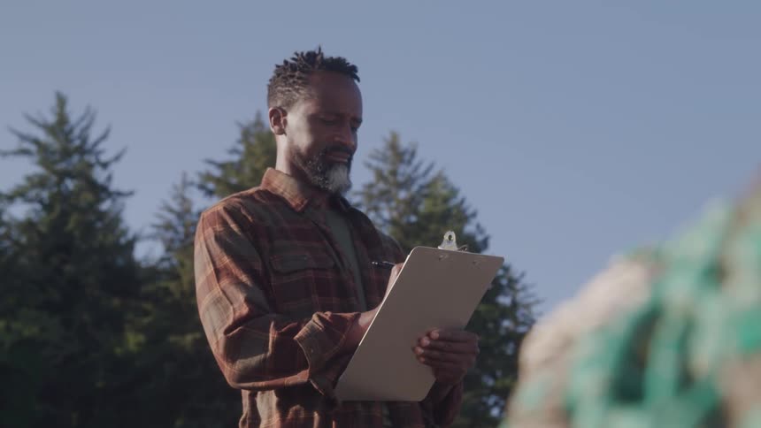 A man outdoors in a rural area writes thoughtfully on a clipboard framed by trees in clear daylight