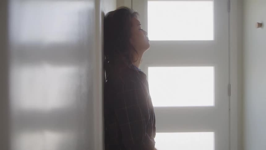 A woman stands and then sits alone by a door looking visibly upset and deep in thought in a quiet sunlit hallway