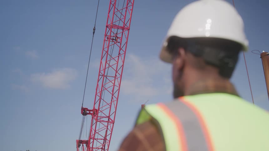 A construction worker in a safety vest and helmet looks up at a red crane against a clear blue sky