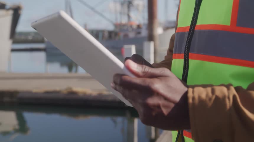 A man typing on a tablet is wearing a safety vest as he stands on the waterfront