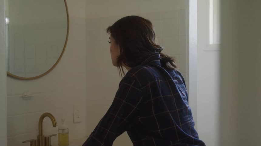 A lady standing inside the bathroom looking in the mirror at dawn