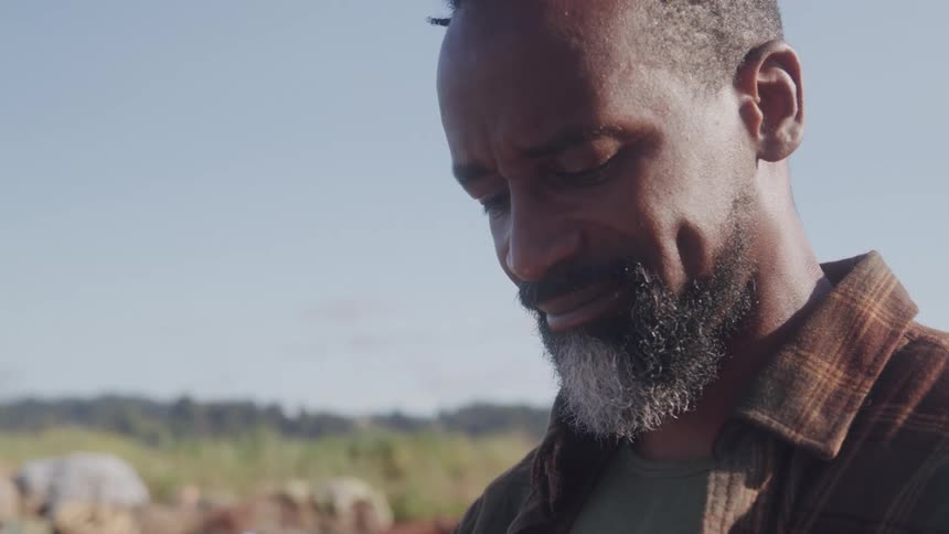 A closeup shot shows a man with a saltandpepper beard looking down and smiling slightly outdoors