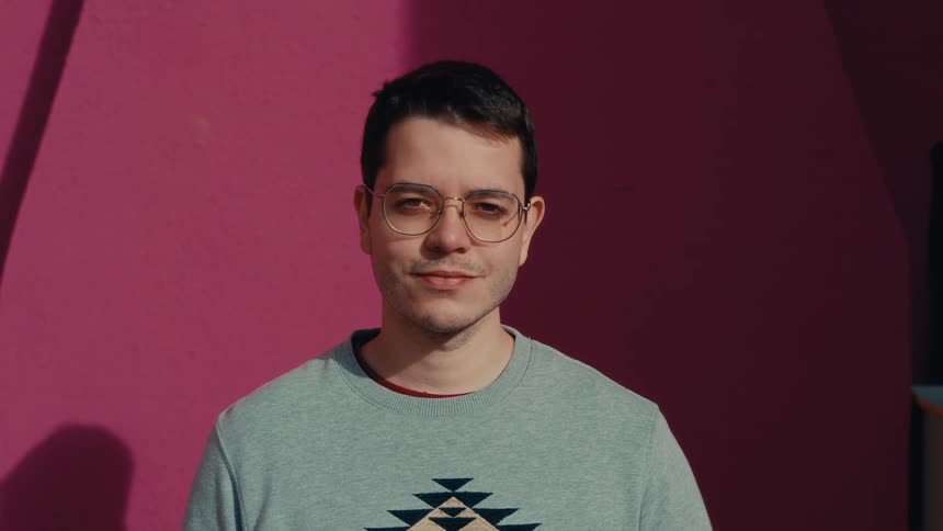 A man stands before a pink wall looking calmly at the camera