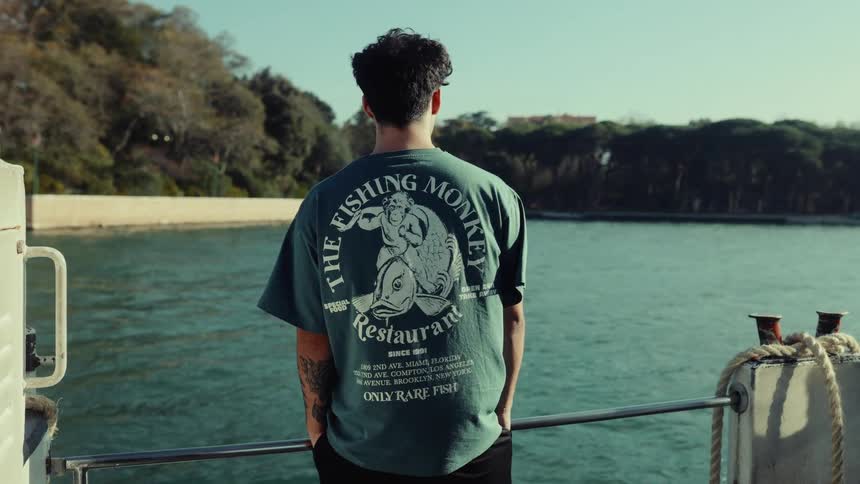 A man stands on a boat deck facing the water wearing a graphic tshirt with a scenic shoreline behind him