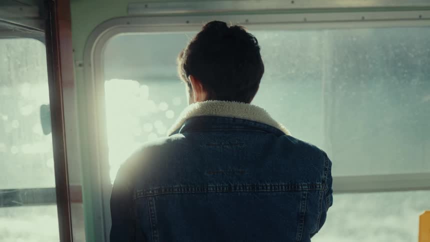 A young man gazes out a boat window his back to the camera sunlight glinting off the water
