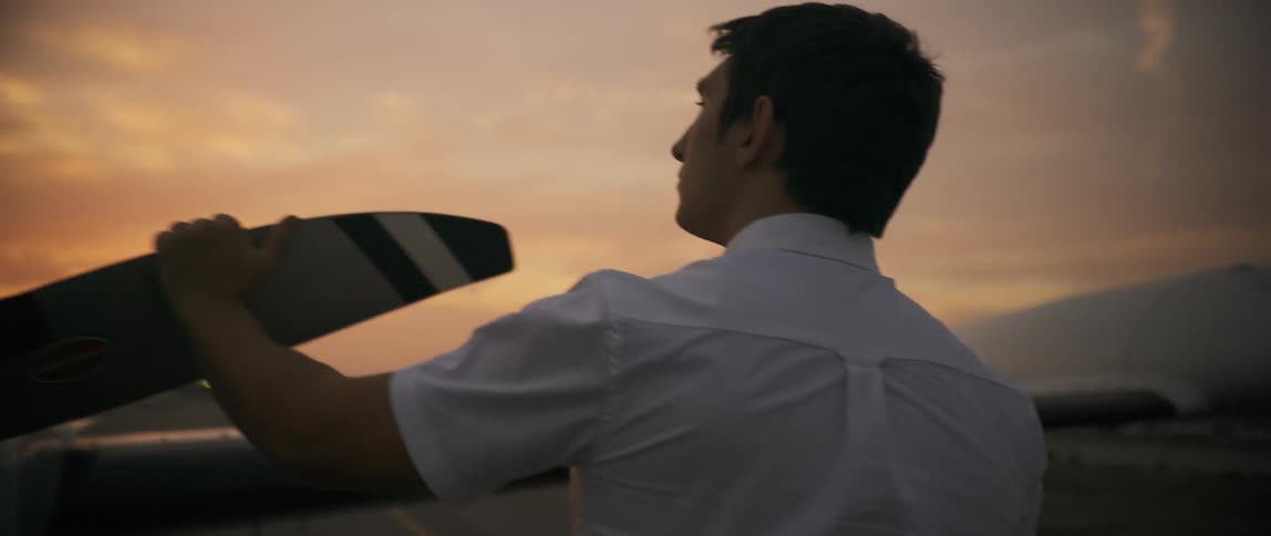 A pilot in a white shirt is inspecting the propeller of a small airplane against a backdrop of a warm dusky sky
