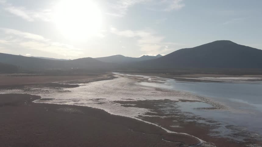 A serene aerial view shows a reflective shoreline at low tide with mountains silhouetted by the setting sun