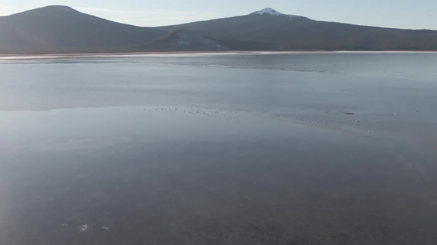 An aerial view of a frozen lake beneath snowcapped mountains under a clear sky