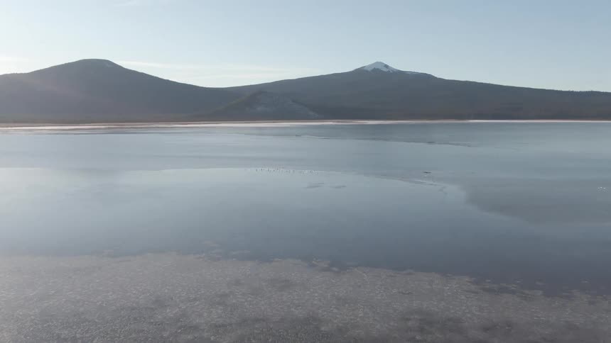 A still lake reflects snowy mountains under a clear daytime sky