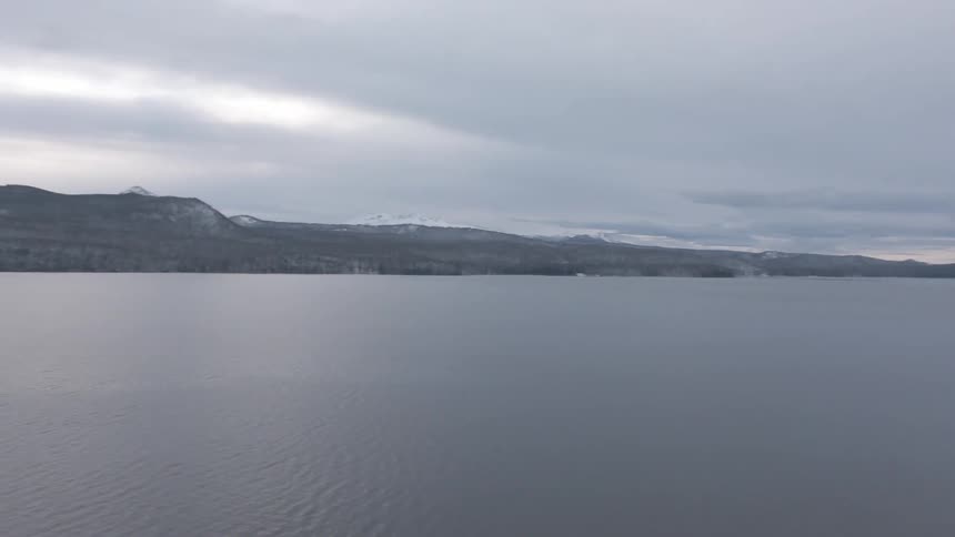 A still lake is framed by evergreen trees and snowy hills beneath a cloudy dusk sky