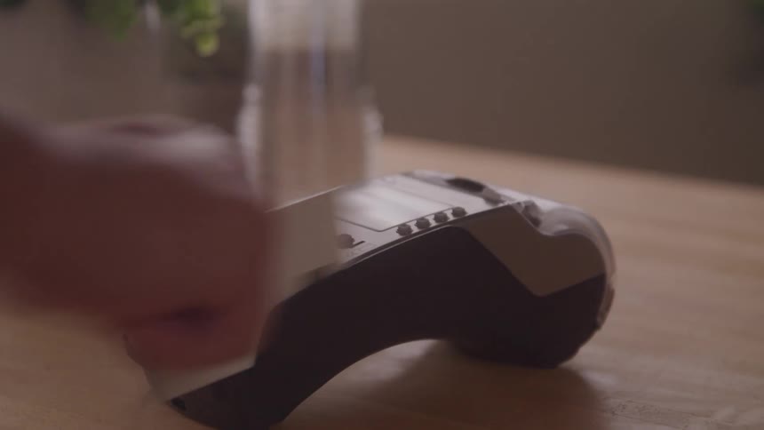A hand swipes a white credit card through a payment terminal resting on a wooden surface