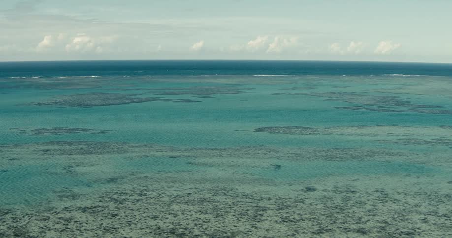 A wide aerial view of shallow coral reefs in turquoise water meeting a deep blue horizon