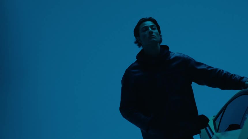 A man wearing a jacket stands beside a car inside a garage with flickering blue light