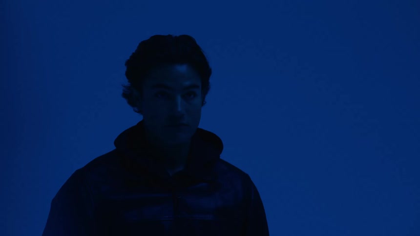 A man stands under deep blue lighting posing calmly for the camera in a minimal moody setting