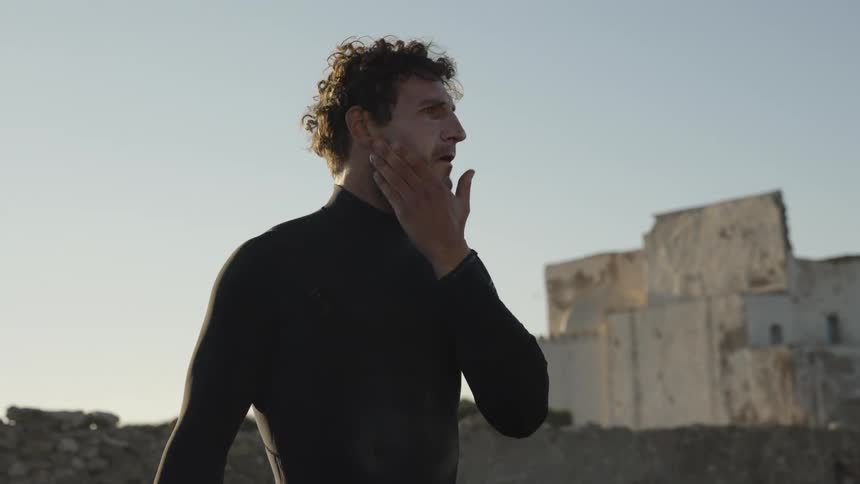 Man wearing a wet suit with a white building in the background wipes his face