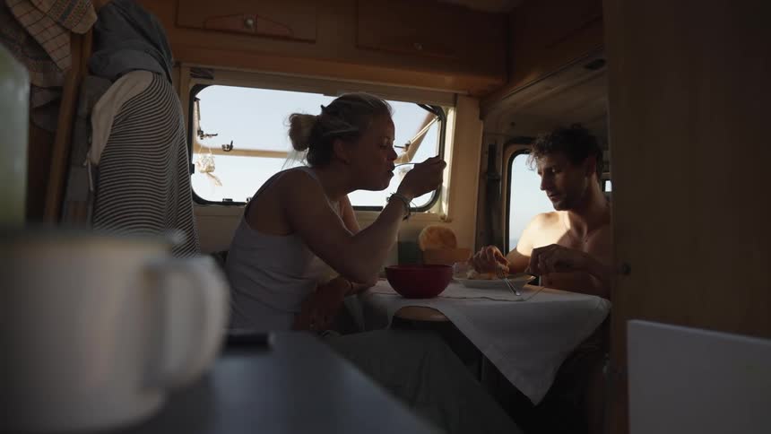 A couple sits inside a camper van sharing a quiet meal together by the window with sunlight streaming in