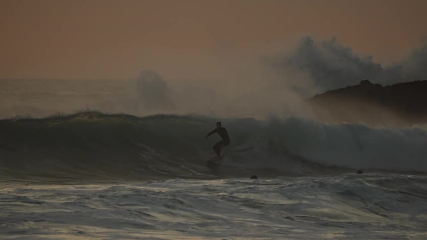 Surfer riding a wave at sunset as others watch from the water near crashing waves and rocks