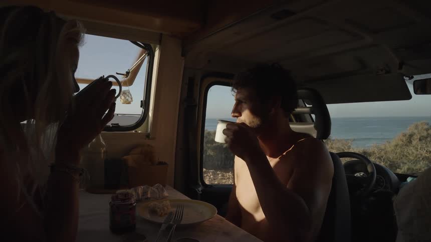 A man and woman enjoy a quiet morning drinking coffee inside a camper van parked near the coast