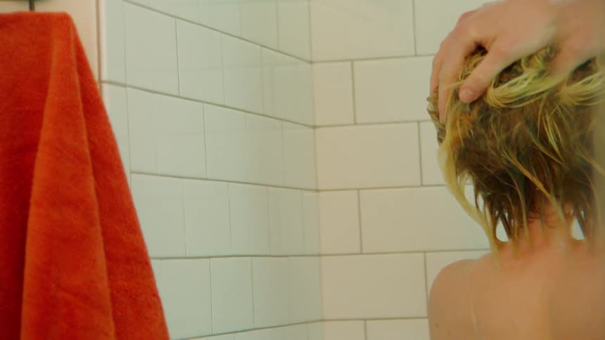 Blonde man rinses his hair while taking a shower in a bathroom tiled in white