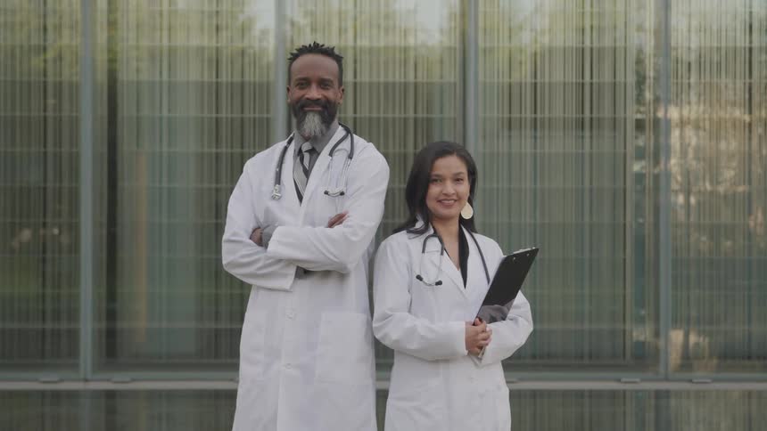 Two smiling doctors in white coats stand posed in front of a building