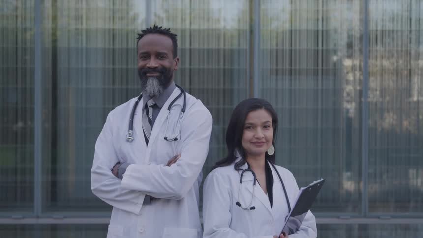 A male and a female doctor both wearing white lab coats with stethoscopes draped around their necks stand outdoors next to each other smiling the female doctor holds a folder and the male doctor who has a saltandpepper beard wears a tie