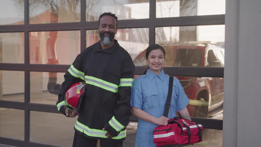 At midday a female paramedic holding a paramedic bag stands next to a male firefighter holding a red helmet both are smiling as they stand in front of a glass building with a fire truck visible through the glass behind them