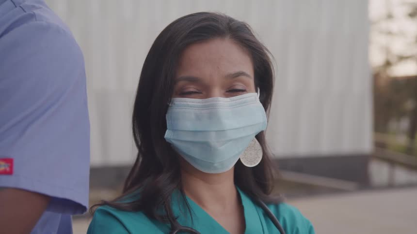 A female paramedic wearing a nose mask stands beside a male outdoors turning her head from the side to face forward