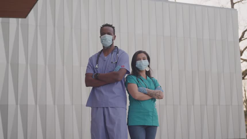 A doctor and nurse stand outside with their arms folded