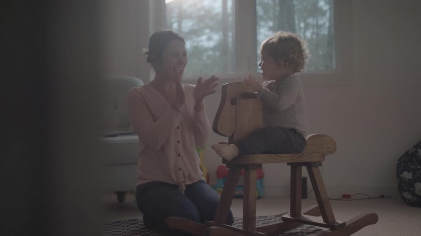 Mother clapping along with her little daughter who is playing on a wooden horse
