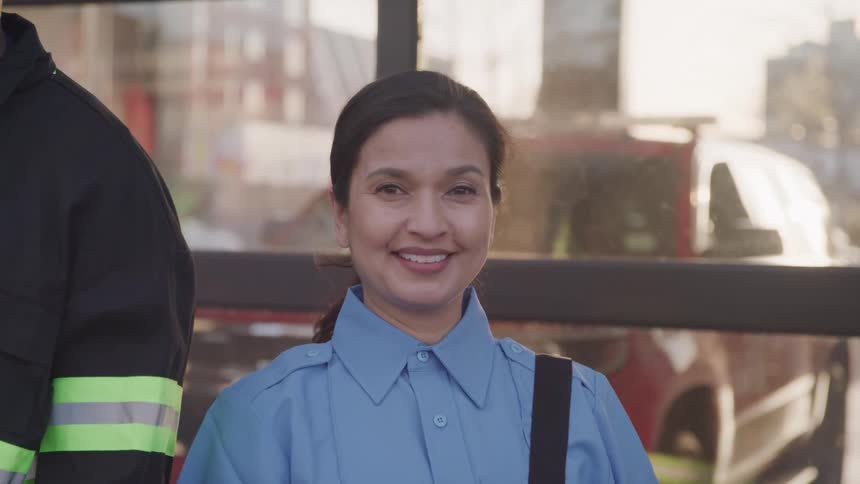 At midday a young lady wearing a strap bag stands next to a firefighter beside a glass wall smiling with a car visible through the glass