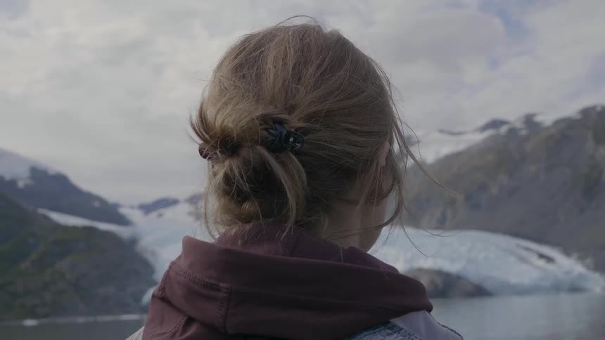 A woman stands by a lake at midday surrounded by snowcovered mountains