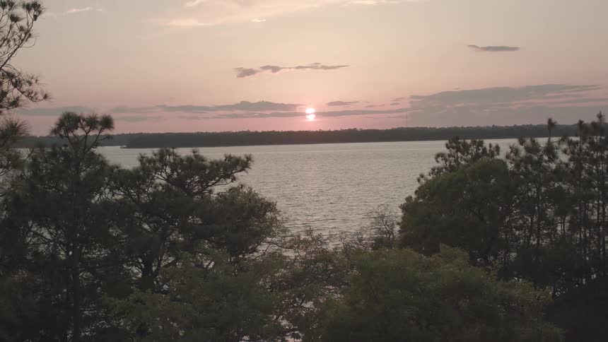 The sun sets over the lakeside casting a warm glow across the water with trees surrounding the lake and reflecting the golden hues of the evening sky