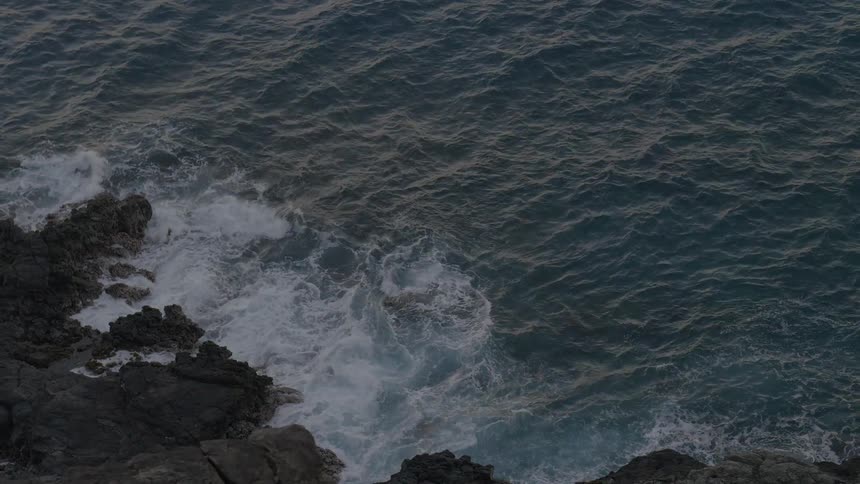 Dark ocean waves repeatedly crash against a rugged rocky shoreline viewed from above