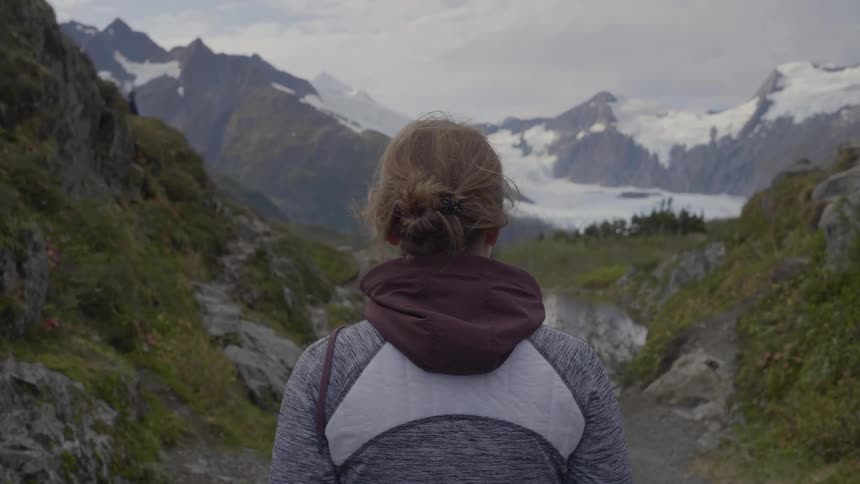 A woman seen from behind hikes along a mountain trail pausing to look at a view of snowcapped peaks a glacier and an alpine lake in the distance