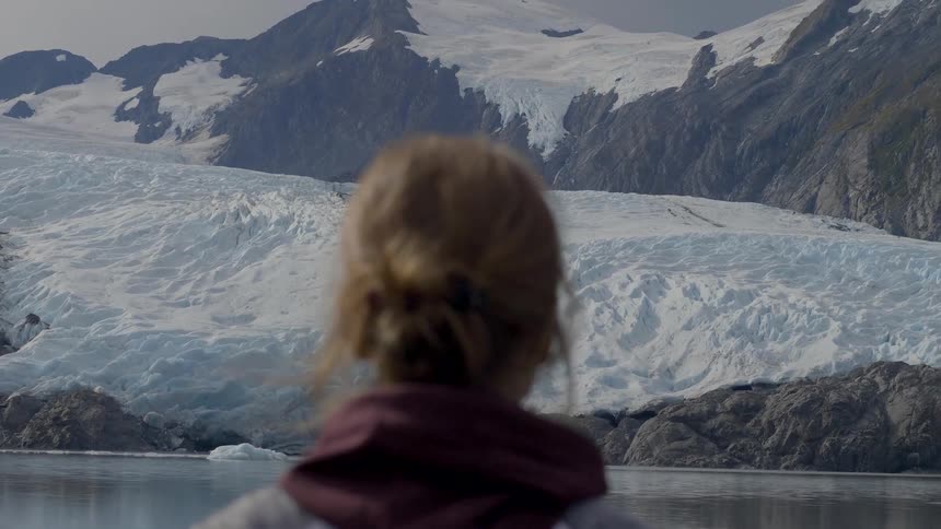 The back of a blonde womans head looking at a glacier at the base of a mountain facing a lake