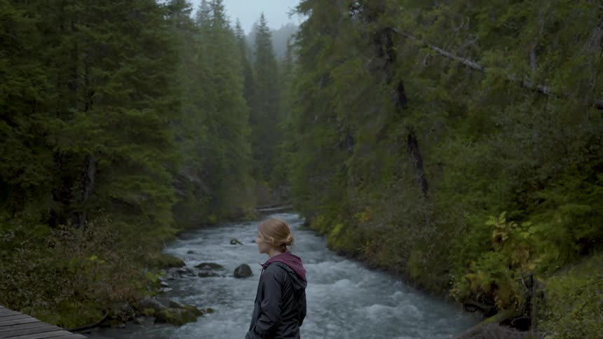 A blonde woman stands on a wooden bridge over a stream flowing through a forested area at midday