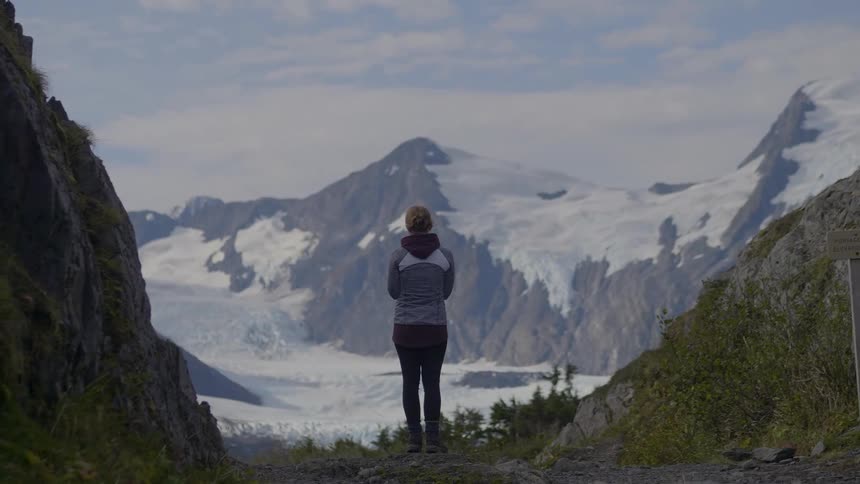A woman stands on a mountain trail gazing at a breathtaking panorama of a vast glacier and snowcovered peaks under a partly cloudy sky