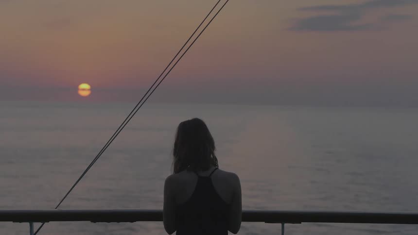 A woman stands on a ships deck her back to the camera watching a serene sunset over the calm ocean horizon