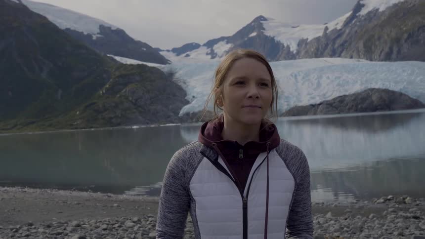 Red haired woman walks away from the coast smiling while the water in the background reflects the nearby snowy mountainous terrain