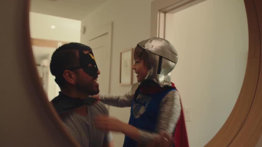 A child in a knight costume smiles while putting on a cape his reflection visible in a mirror beside an adult wearing a superhero mask and cape