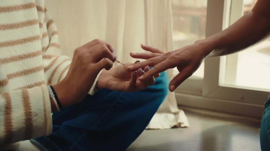 A young teenager sits indoors next to a window at midday laughing as she carefully applies nail polish to another girls right fingers with sunlight filtering through the glass