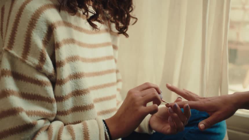 A teenage girl is talking while painting the nails of a person in front of her