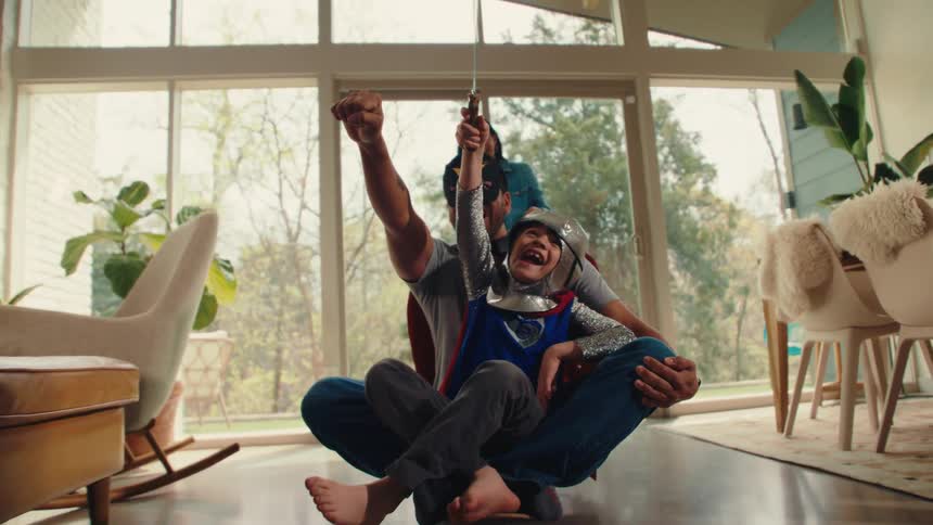 A man and child sit on a skateboard wearing superhero costumes and masks the child holding a sword both raising their hands with joy as the mother pushes the skateboard forward in the living room at midday