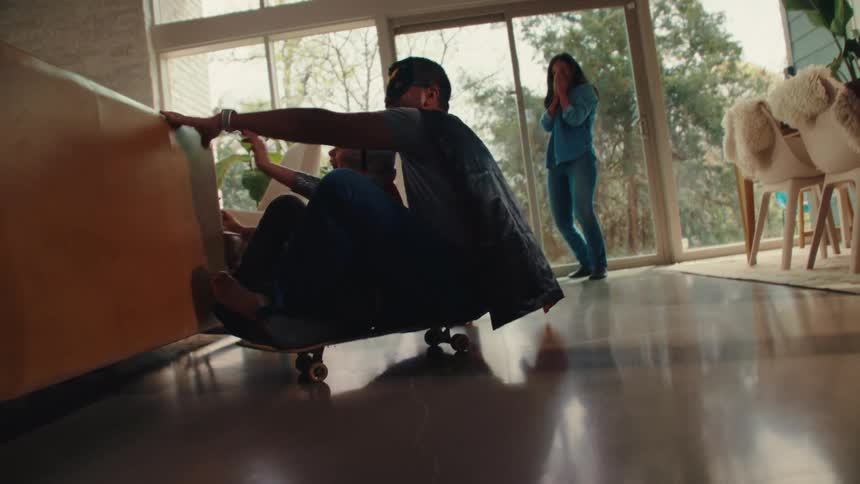 Father and child in costumes ride a skateboard indoors as a woman reacts behind them